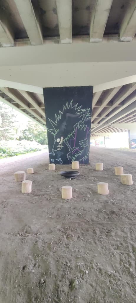 Vuurschaal met logbanken rond een graffiti-pilaar onder het viaduct